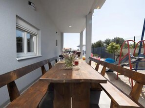 Enjoy meals al fresco in this outdoor dining area with a long table and benches, surrounded by a playground and lush greenery.