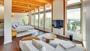 Ein geräumiges Zimmer mit einem großen Bett, einer Sitzecke und einem Fernseher. Mehrere Fenster bieten Blick auf Bäume und einen See.