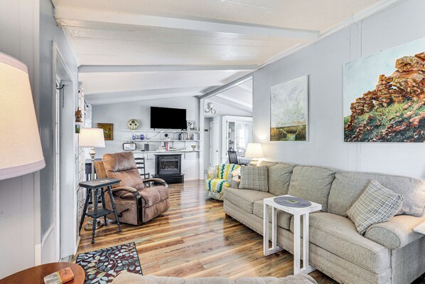 A cozy living room with a sofa, armchair, and a small table. A fireplace is visible in the background, and there are framed pictures on the wall. - Lake Marion Canal Home: Dock, Yard, Porch (Summerton)