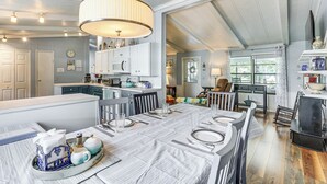 Une salle à manger spacieuse avec une grande table dressée pour six personnes, une cuisine en arrière-plan et une vue sur l’extérieur par une fenêtre.