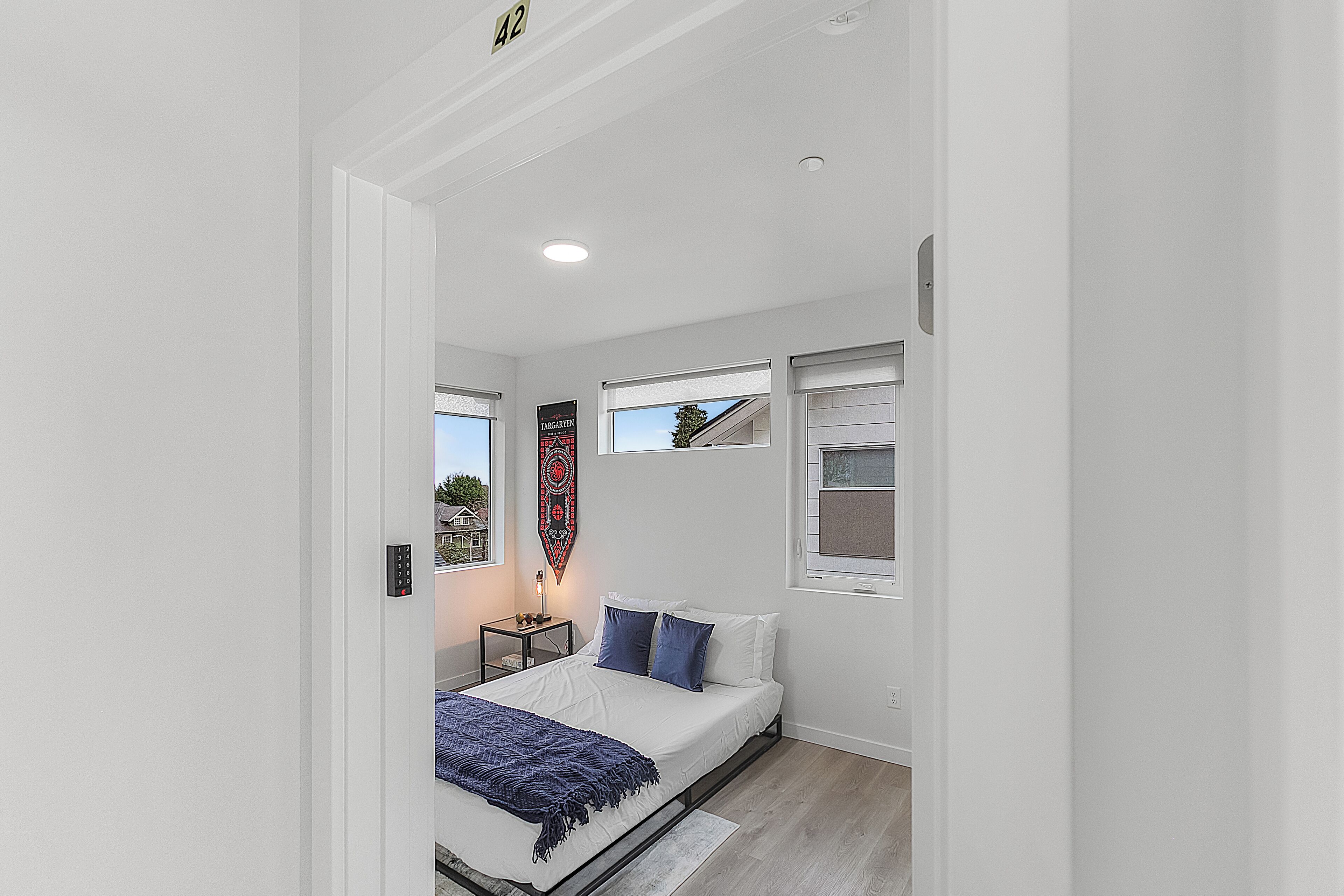 Une chambre moderne avec un lit, deux fenêtres, une table de chevet et un luminaire fixé au mur.