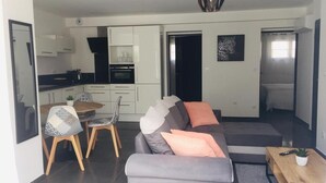 Una sala moderna con un sofá gris, una mesa de comedor con sillas, y una cocina con armarios blancos y encimera negra.