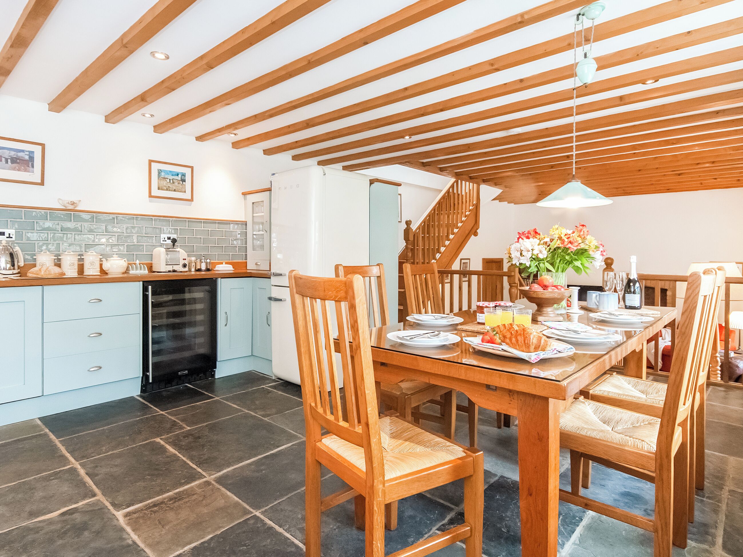 A dining area with a wooden table set for four, surrounded by chairs. A kitchen with light blue cabinets and a black refrigerator is visible in the background.