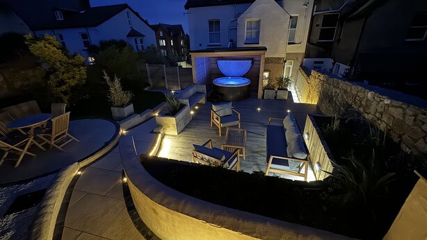 A well-lit outdoor patio area with a hot tub, seating, and a stone wall.