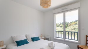 Une chambre avec un grand lit, du linge de lit blanc, une vue sur la verdure et une lampe en osier.
