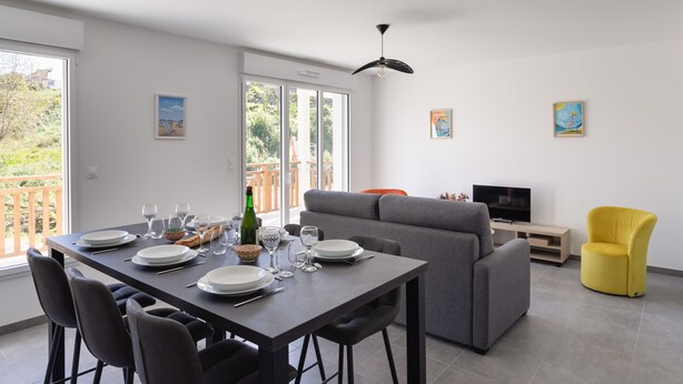 Un coin repas avec une table dressée pour quatre personnes, un canapé gris, un fauteuil jaune et un téléviseur posé sur un meuble en bois.