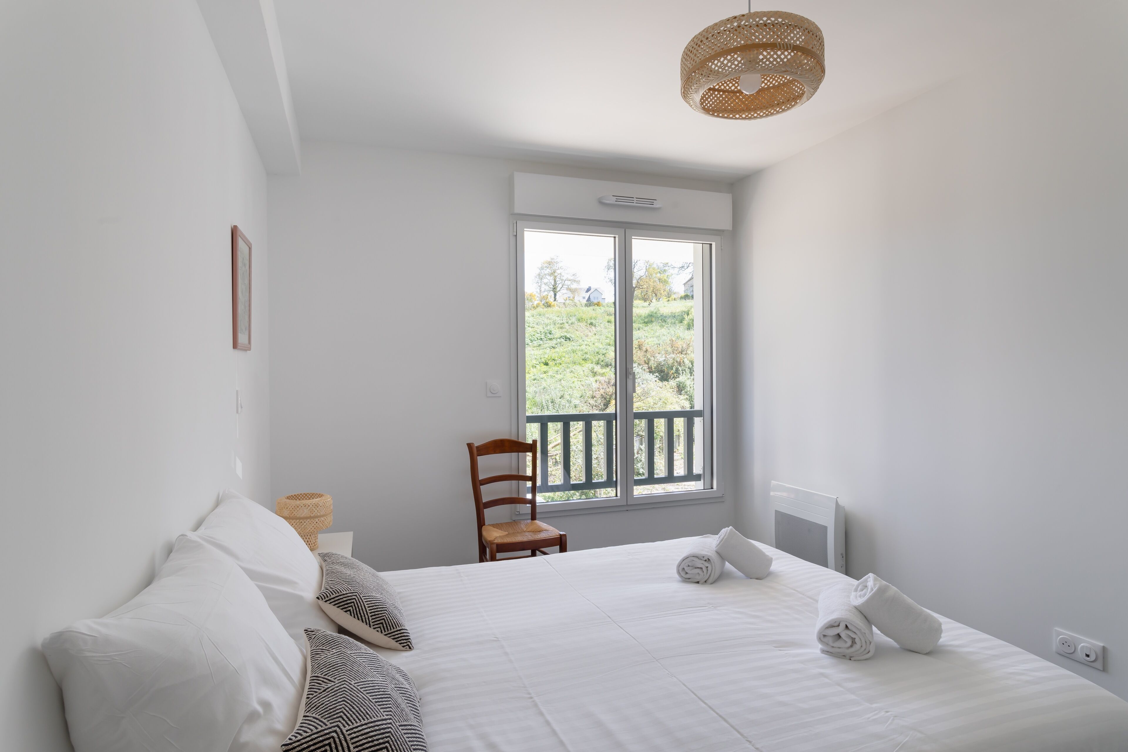 Une chambre avec un grand lit, une chaise, une fenêtre donnant sur un paysage verdoyant et un luminaire au plafond.