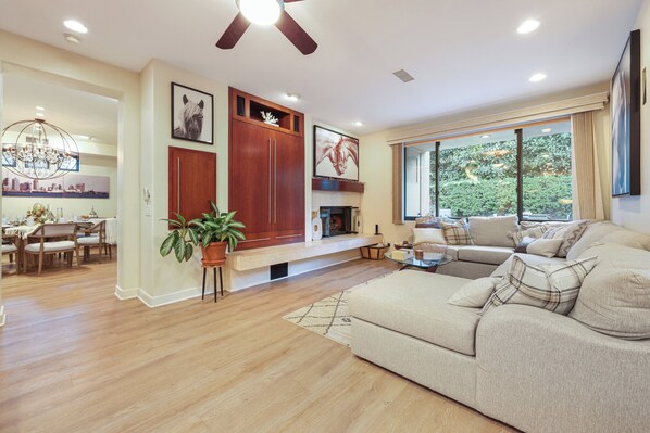 A spacious living room with a large sofa, a fireplace, a dining area with wooden furniture, and a view of greenery through large windows. - Sunset Beach House in Heart of La Jolla Shores (San Diego)