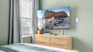 Une chambre à coucher avec un lit, un téléviseur fixé sur une commode en bois et une fenêtre avec des rideaux.