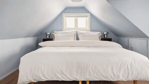 Ein Schlafzimmer mit schrägen Wänden, einem großen Bett mit weißer Bettwäsche, zwei Nachttischen und einem Fenster mit weißen Rollläden.