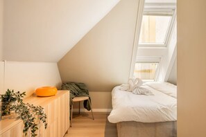 A bedroom with a sloped ceiling, a bed with white bedding, a small wooden dresser, and a window with blinds.