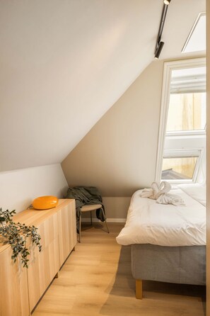 A bedroom with a sloped ceiling, a bed with white bedding, a wooden dresser, and a window with blinds.