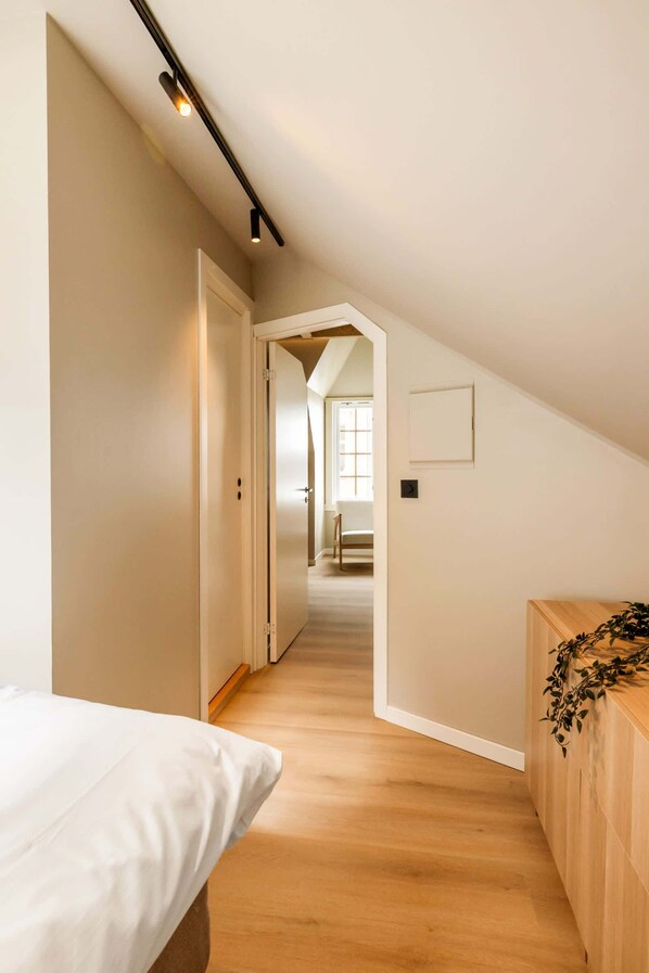 A narrow hallway with wooden flooring, a bed on the left, and a wooden bench on the right.