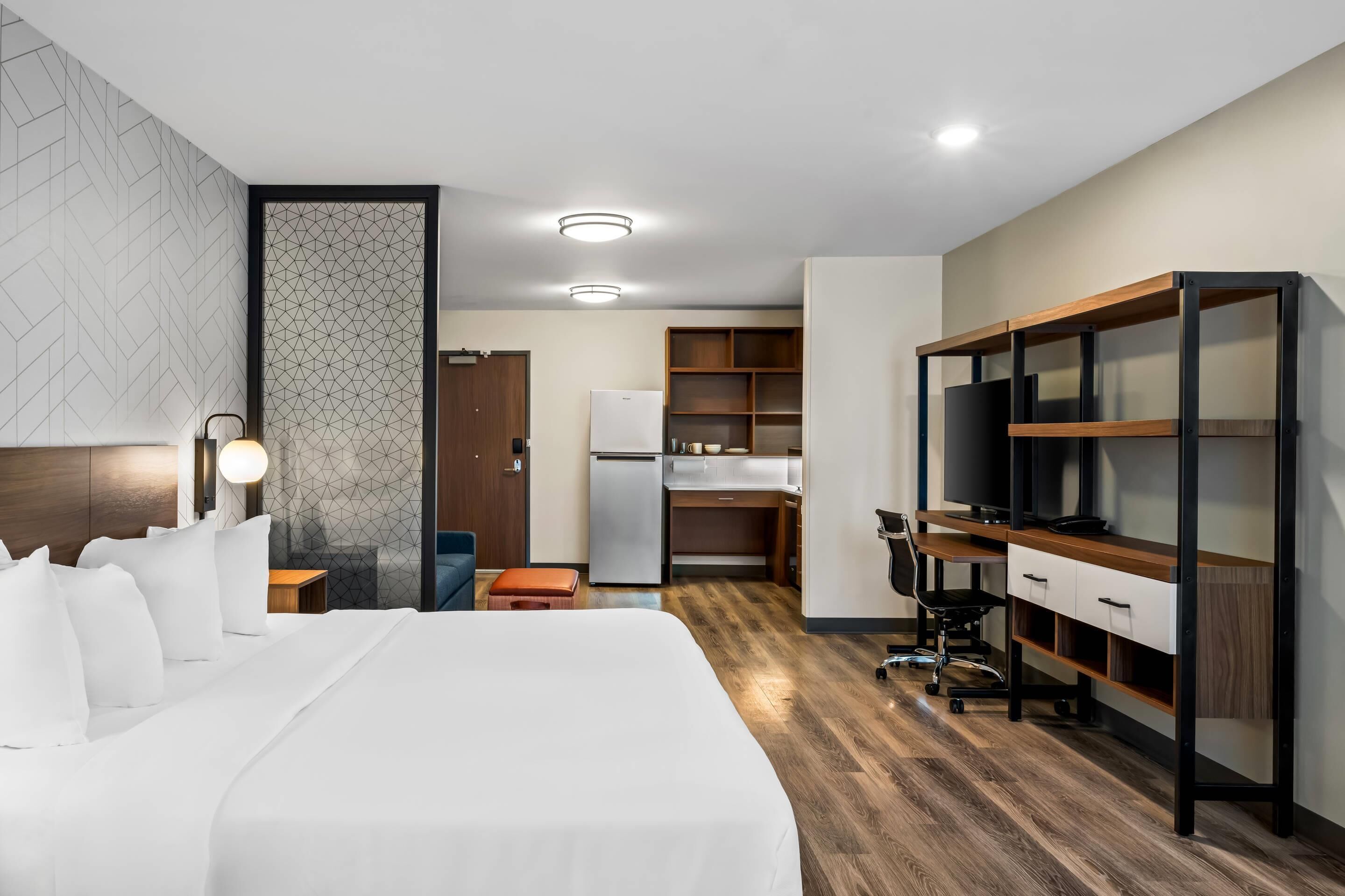 A hotel room with a large bed, a desk, a TV, and a kitchenette.