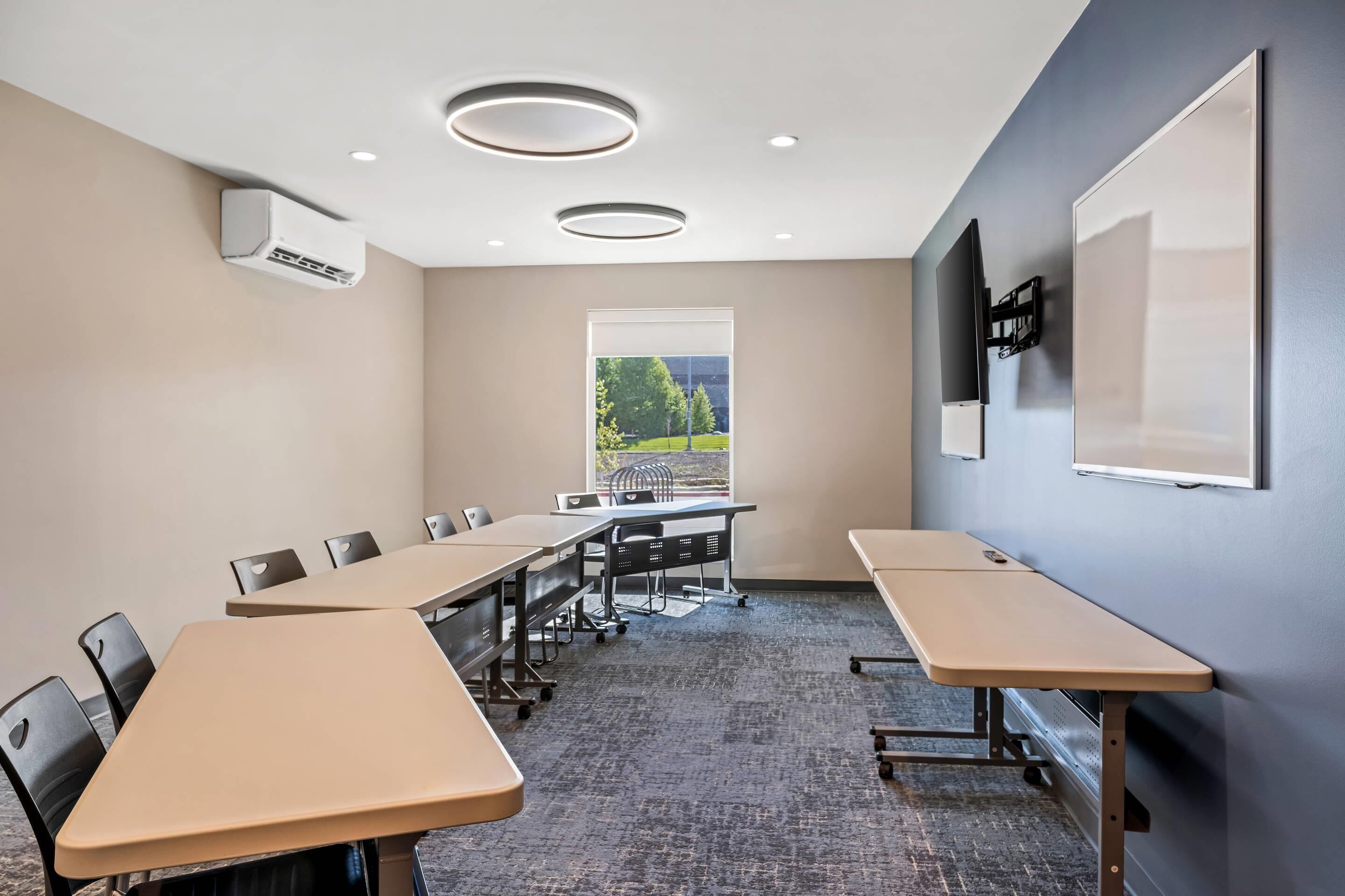 A conference room with a large table, chairs, a projector screen, and a window with a view of trees and a building.