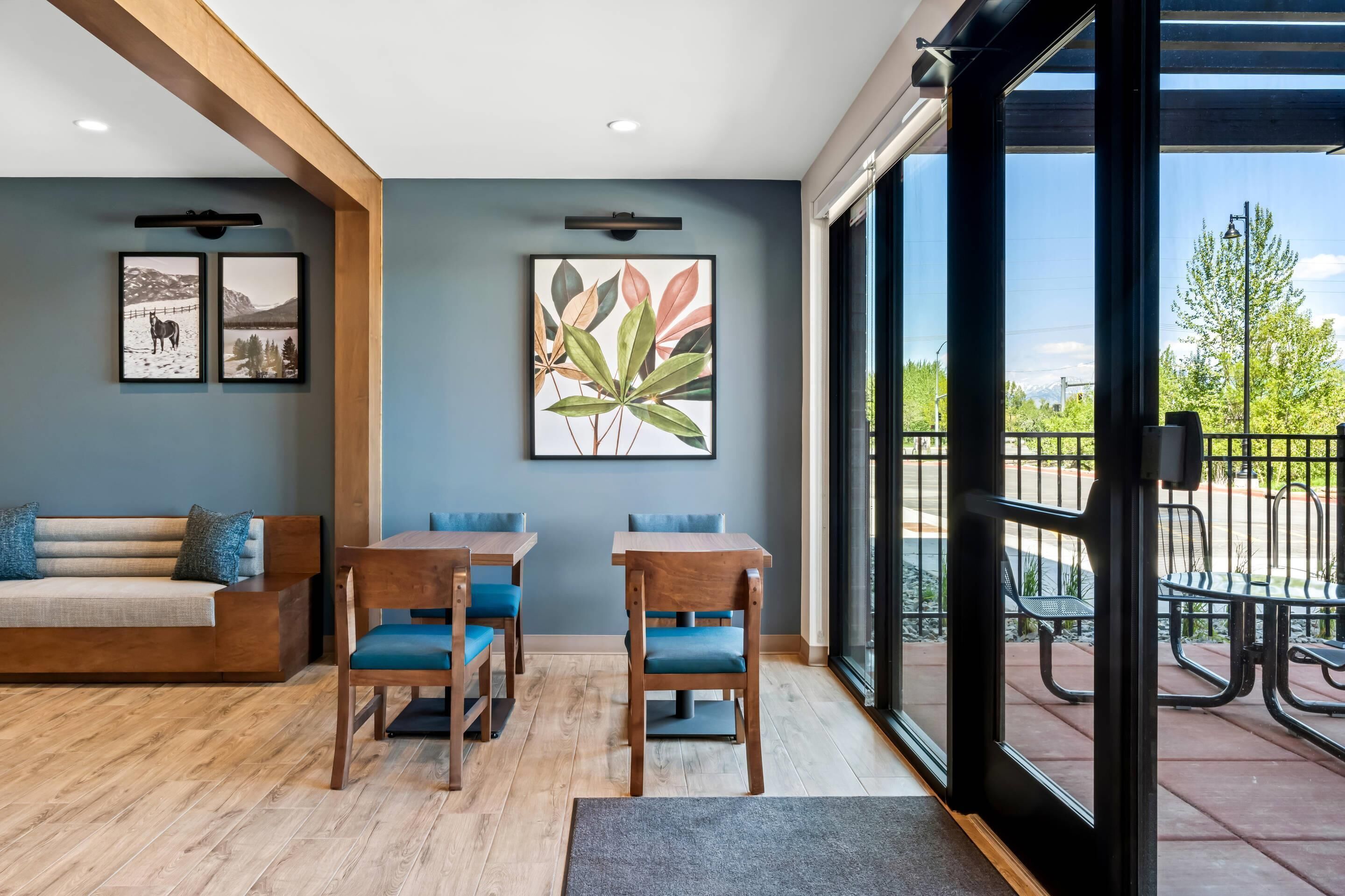 A modern interior with a wooden sofa, two tables with blue chairs, and a large window with a view of a patio area.