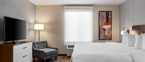 Una camera d'albergo con televisione a schermo piatto, una poltrona grigia, un letto grande con lenzuola bianche, una lampada da terra e un quadro appeso al muro.