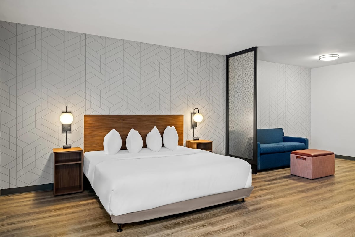 A modern hotel room with a large bed, two bedside tables with lamps, a blue sofa, and a small wooden stool.