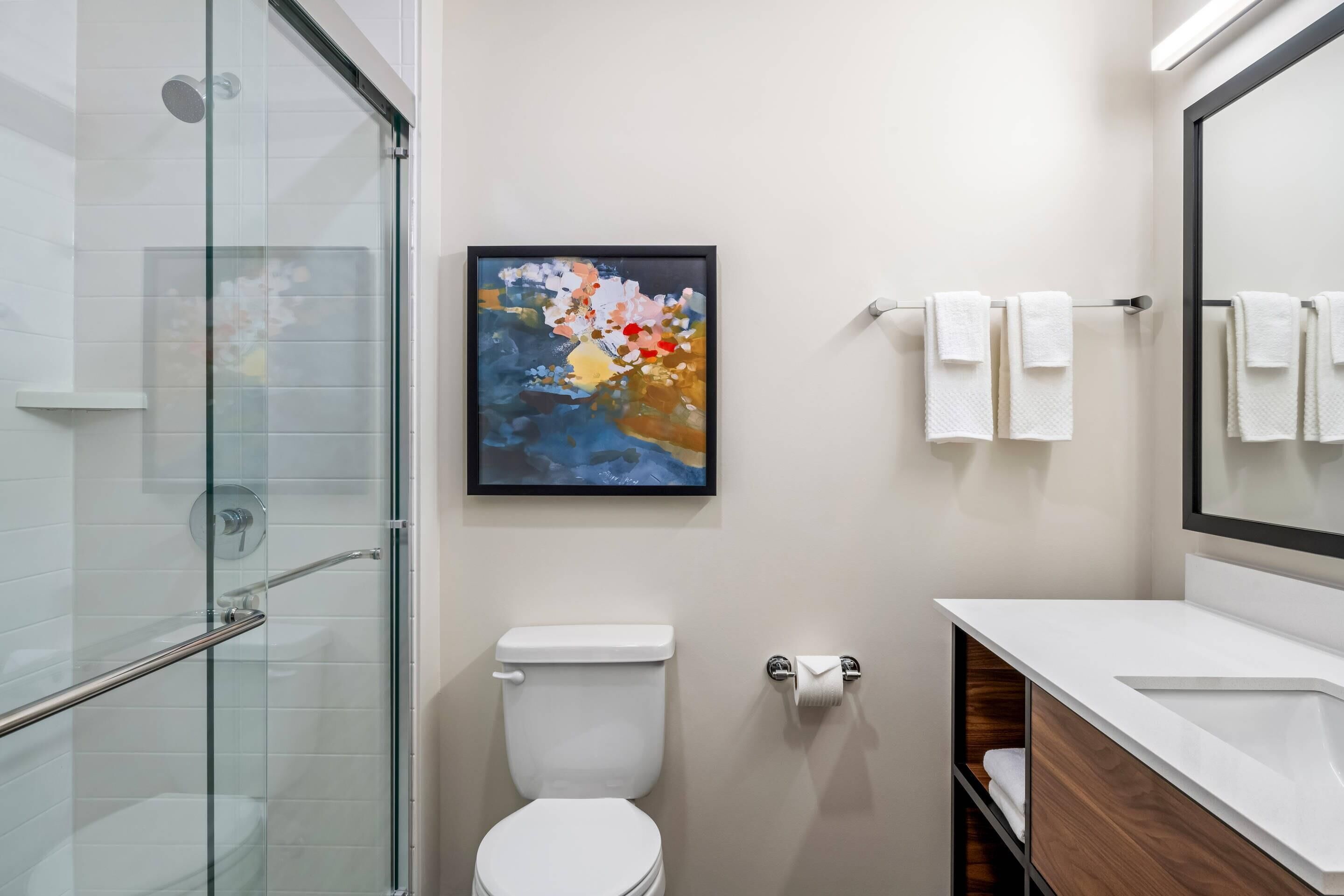 A modern bathroom with a glass shower enclosure, a white toilet, a framed abstract painting, and a white sink with a wooden vanity.