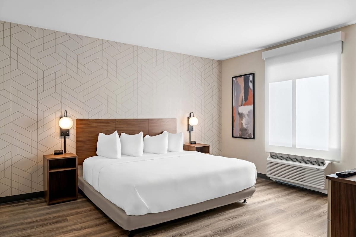 A hotel room with a large bed, two bedside tables with lamps, a wooden headboard, a geometric-patterned wall, a large window with blinds, and a wall-mounted air conditioner.
