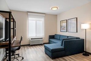 A modern hotel room with a wooden desk, a blue sofa, a standing lamp, and framed artwork on the wall. - Everhome Suites Bozeman (Bozeman)