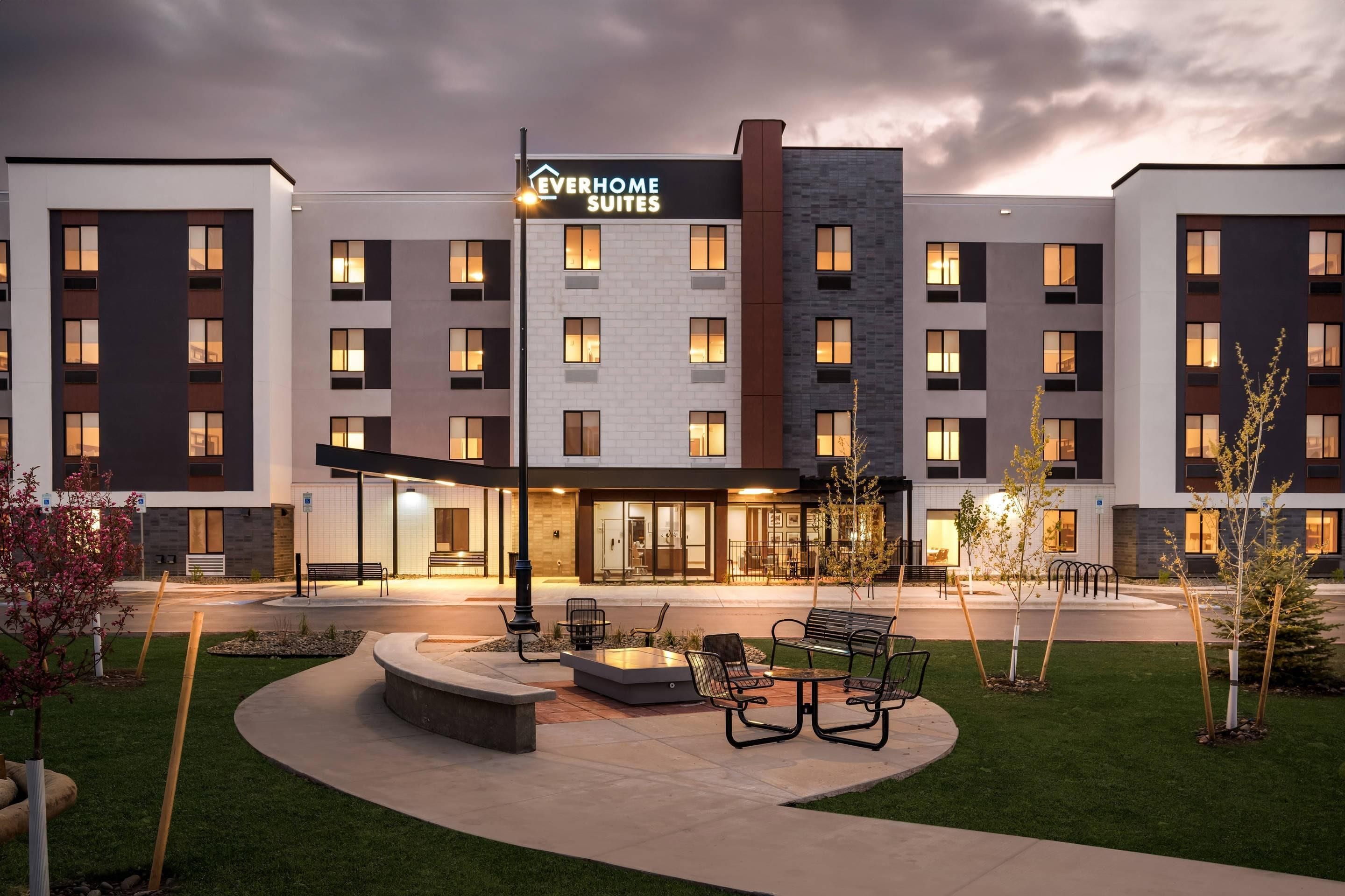 Un hotel moderno con varios pisos, mobiliario exterior y una entrada bien iluminada.