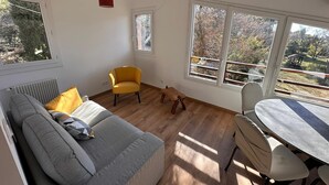 Una sala moderna con un sofá gris, un sillón amarillo y una mesa de comedor con sillas. Amplias ventanas ofrecen vistas a árboles y a un balcón.