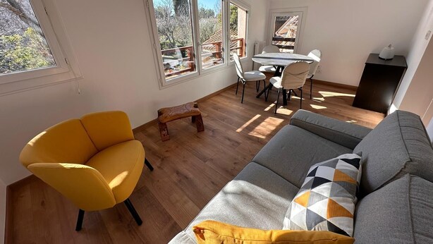 Una sala de estar moderna con un sillón amarillo, un sofá gris con cojines estampados y un banco de madera. Hay una mesa de comedor con sillas y una ventana con vistas a árboles y un edificio.