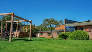 Un ensemble de balançoires en bois installé dans un espace verdoyant, avec un bâtiment portant l’inscription « RÉCEPTION » et une enseigne indiquant « BAR » en arrière-plan.