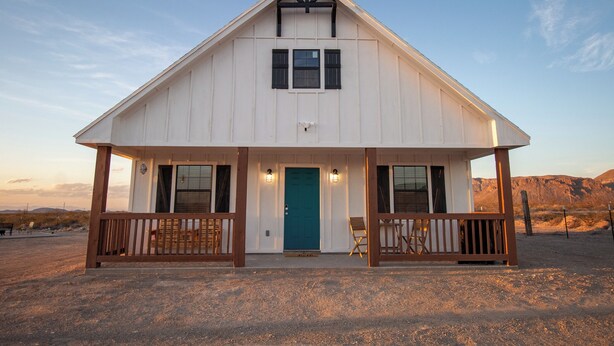 A white building with a gabled roof, a front porch with wooden railings, and a blue door.