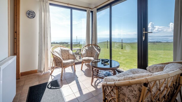 A room with wicker furniture, a glass table, and a view of a grassy field and sea. - Barnstable - UK46888 (Shannochie)