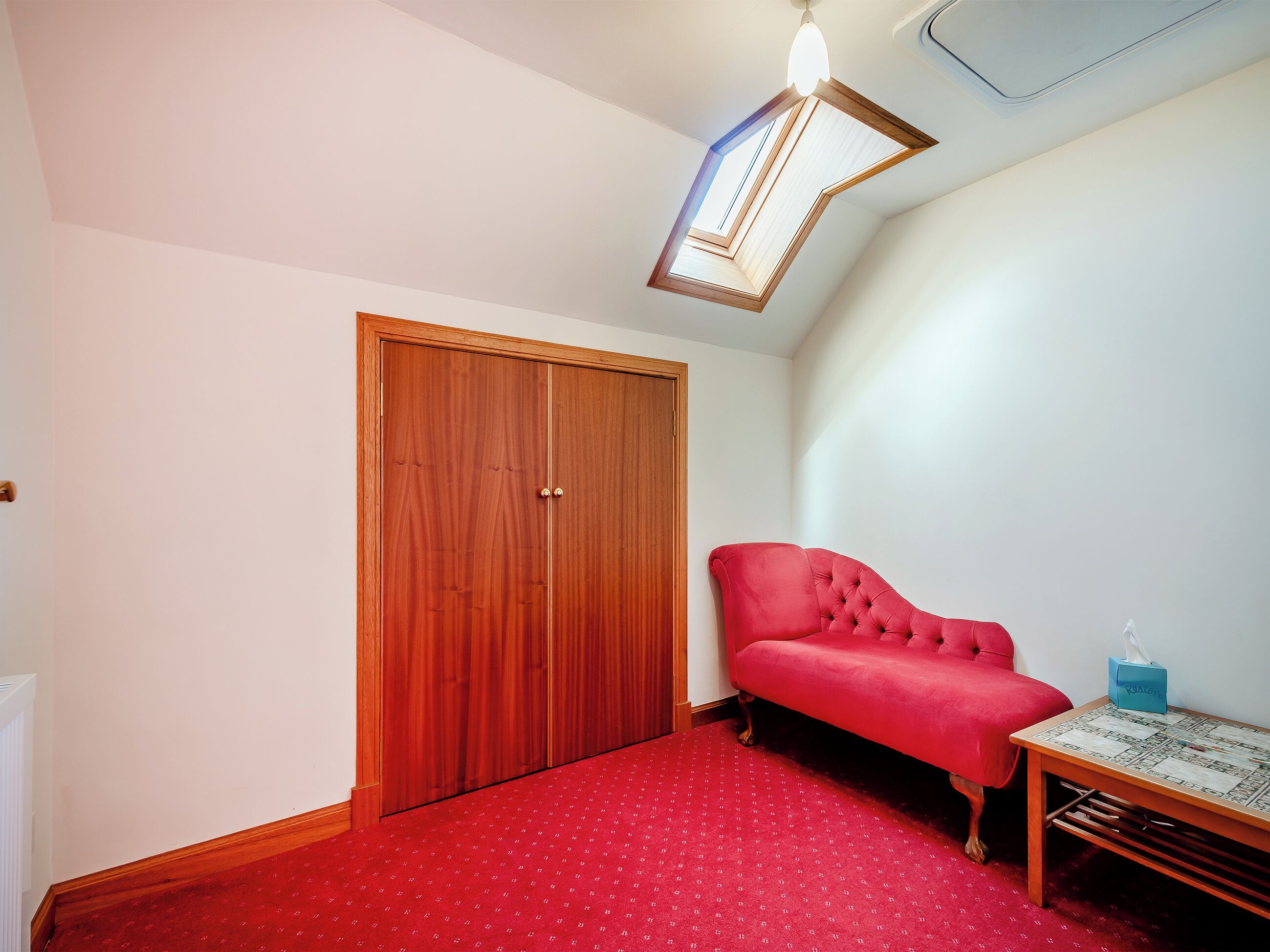 A room with a red carpet, a wooden wardrobe, a red sofa, and a small table with a tissue box.