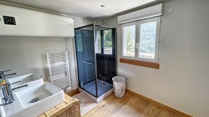 A bathroom with a white sink, a glass shower enclosure, a white radiator, a window with a view of greenery, and a wooden floor. - Chalet Familial Dans les Monts D'ardèche (La Souche)