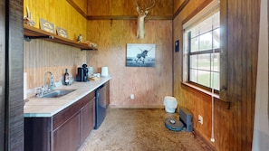 Une cuisine rustique aux murs en bois, ornée d’une tête de cerf fixée au mur, avec une machine à café, des verres à vin et une fenêtre offrant une vue agréable.