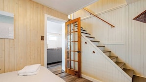 Een slaapkamer met een bed, een houten deur die leidt naar een trap, en zicht op een andere kamer.