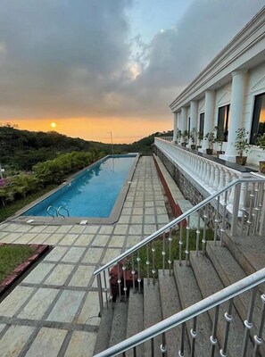 A swimming pool with a stone deck, a staircase leading to a balcony with columns, and a sunset in the background.