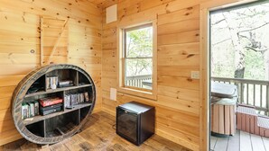 Interior de una cabaña de madera con una estantería, un pequeño electrodoméstico y una ventana con vista a árboles.