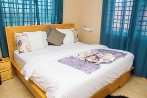 Ein ordentlich gemachtes Bett mit weißen Leinen und einer violetten Decke, ein Holz-Kopfbrett und ein Nachttisch mit Schubladen.