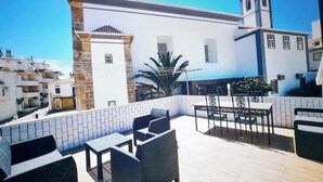 Une terrasse sur le toit, dotée d’un espace extérieur aménagé et offrant une vue sur un bâtiment blanc surmonté d’un clocher.