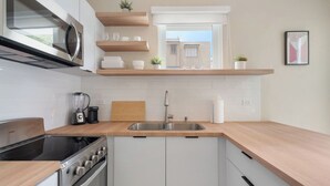 A modern kitchen with a built-in stove, microwave, and double sink. There are wooden shelves above the counter and a window providing a view of buildings outside.