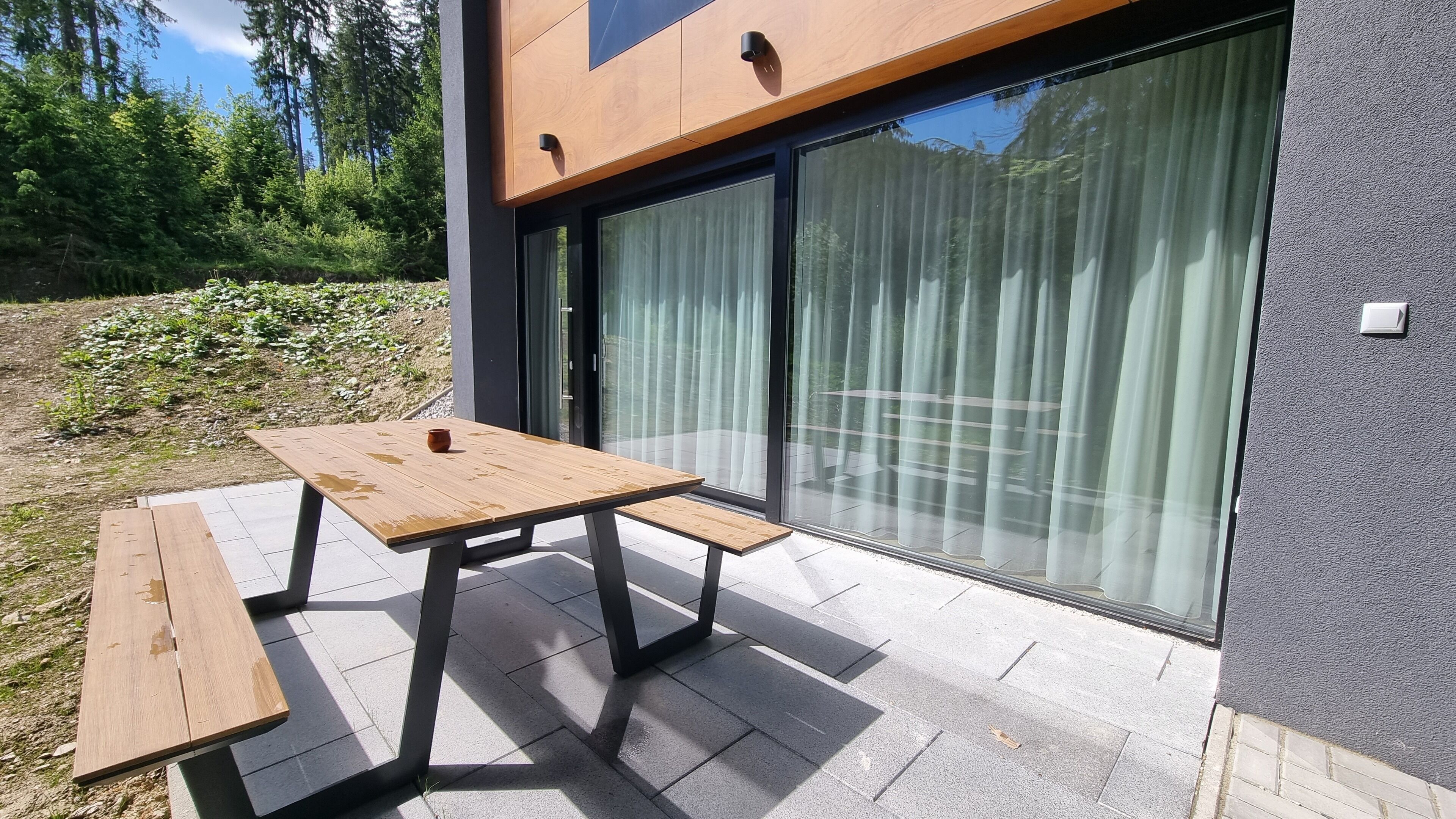 An outdoor patio with a wooden table and benches, a sliding glass door, and a grey textured wall.