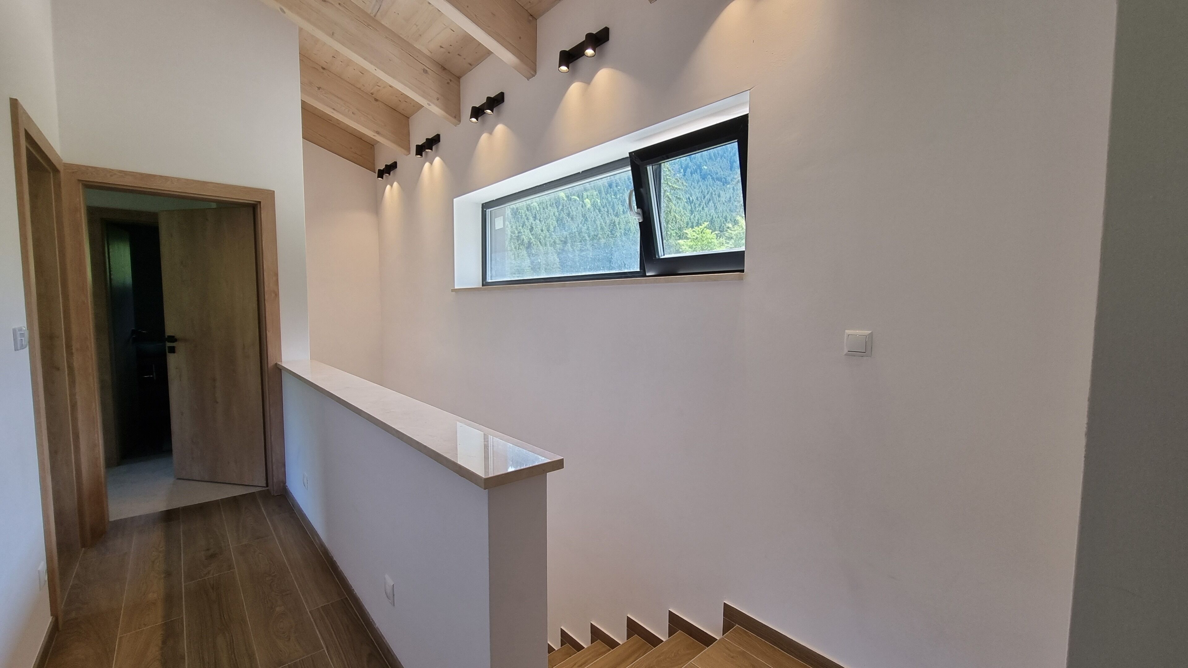A hallway with a wooden floor, a white wall, and a window with a view of trees.