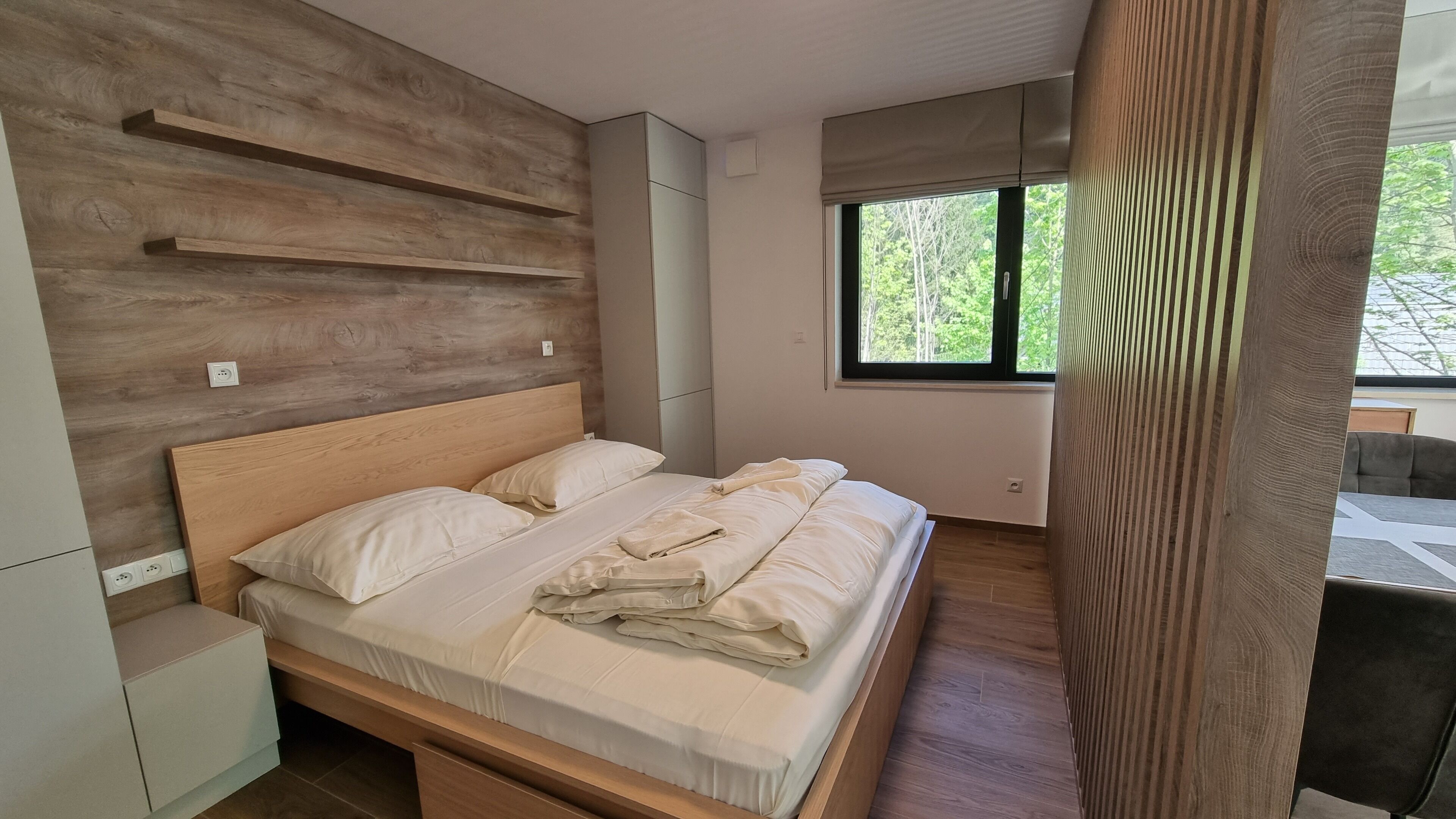 A modern bedroom with a wooden headboard, a bed with white linens, and a window providing a view of greenery outside.