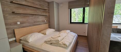 A modern bedroom with a wooden headboard, a bed with white linens, and a window providing a view of greenery outside.