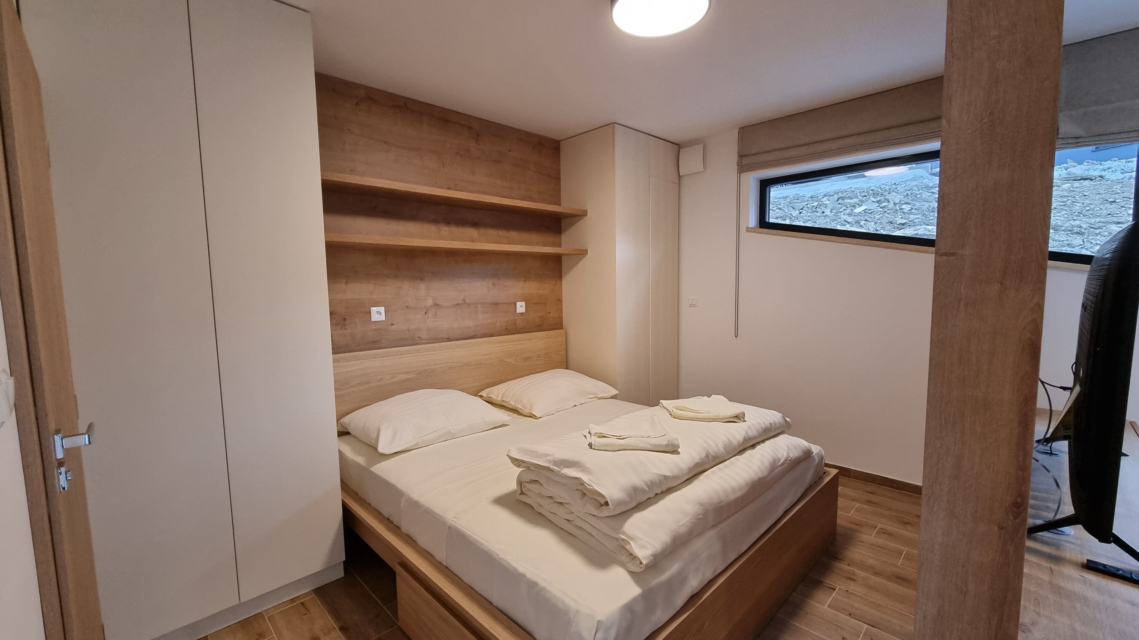 A bedroom with a wooden bed, a wardrobe, and a window with a view of a rocky landscape.