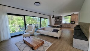 A modern living room with a sofa, armchair, and coffee table, a dining area with chairs, and a kitchen with wooden cabinets and a large window. - Chalets Hrabovo (Ruzomberok)