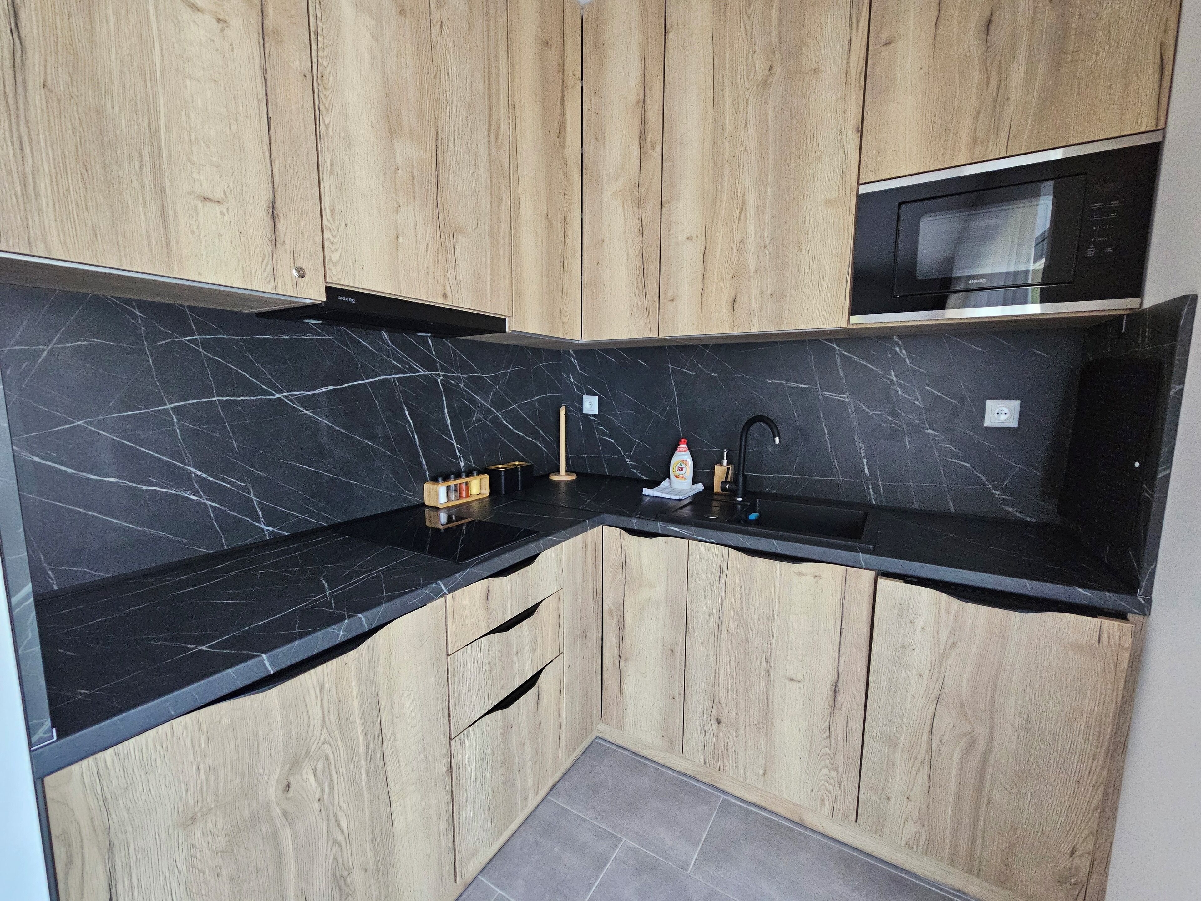 A modern kitchen with black marble countertops, wooden cabinets, a black sink, and a microwave.