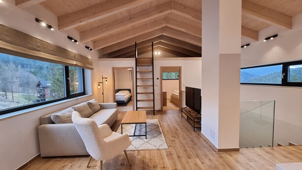 A spacious living room with a high ceiling, wooden beams, a large window, a sofa, a coffee table, and a TV. - Chalets Hrabovo (Ruzomberok)