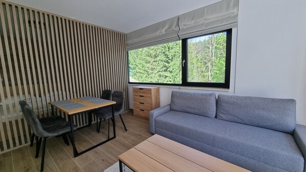 Un espacio habitable pequeño con una mesa de madera, dos sillas, un sofá gris y una ventana con vistas a árboles.