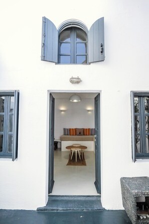 A white building with a central doorway leading to a room with a sofa, two round tables, and a ceiling light.